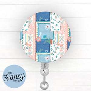 May include: A round badge reel with a patchwork design featuring blue, pink, and white floral patterns. The reel has a silver clip and a clear plastic strap. The text "The Sidney Studio" is on a light blue circle.