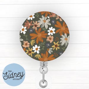 May include: A round floral badge reel featuring a pattern of orange, white, and yellow flowers on a dark green background. The reel has a clear plastic strap and a metal clip. The text "The Sidney Studio" is on a light blue circle.