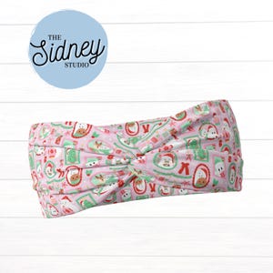 May include: A pink headband with a Christmas-themed pattern featuring Santa Claus, snowmen, and festive frames. The headband has a knotted design and the text "The Sidney Studio" in a light blue circle.