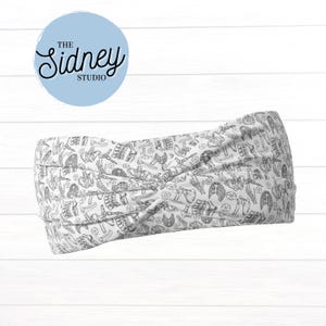 May include: White headband with a black and white anatomical print. The headband features a twisted knot design. The image includes a light blue circle with the text "The Sidney Studio" in a script font.