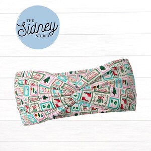 May include: A cream-colored headband with a festive Christmas pattern. The fabric features a repeating design of Christmas trees, boots, and the words "Merry Christmas" in red, green, and teal. The headband is knotted at the center.