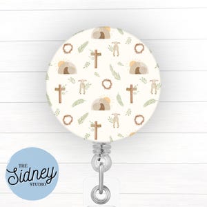 May include: A round, cream-colored badge reel with an Easter-themed pattern. The design includes crosses, lambs, wreaths, and a depiction of the empty tomb. The text "The Sidney Studio" is on a light blue circle.