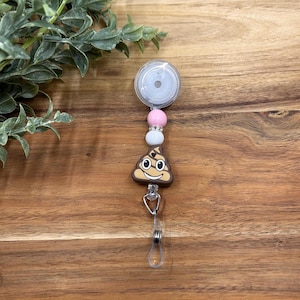 May include: A brown poop emoji charm with a smiling face on a retractable badge reel with pink and white beads.