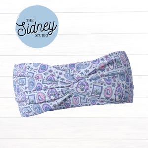 May include: A light blue headband with a repeating pattern of snowmen, hats, and other winter-themed illustrations in shades of purple and pink. The headband has a knotted design and features the text "The Sidney Studio" in a light blue circle.