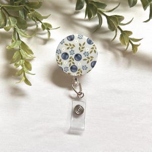 May include: A round retractable badge reel with a blueberry pattern. The design features blueberries, white flowers, and green leaves on a light blue background. The reel has a clear plastic strap and a metal clip.