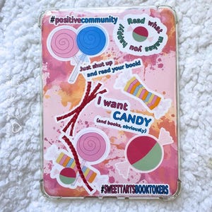 May include: A clear protective case for an e-reader with a pink and orange watercolor background. The case is decorated with candy and book-themed stickers, including the phrases "I want CANDY" and "Read what makes you happy".