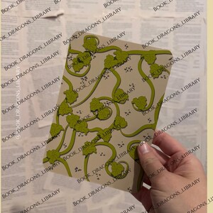 Pu&ograve; includere: Una carta rettangolare con sfondo beige e motivo a vite verde con foglie. La carta &egrave; tenuta in mano su uno sfondo di pagine di vecchi libri. Il testo "BOOK_DRAGONS_LIBRARY" &egrave; stampato sulla carta e sullo sfondo.