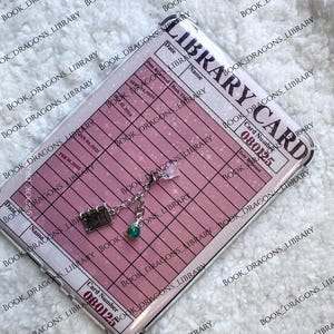 May include: A clear phone case featuring a pink library card design with the text "LIBRARY CARD" and "BOOK DRAGONS LIBRARY". A charm with a book, a green gem, and a clear crystal hangs from the case. The case has a glittery effect.