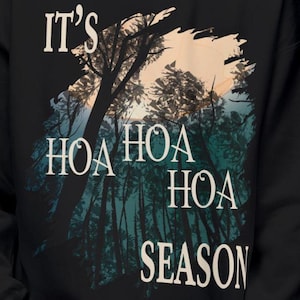 May include: A black sweatshirt featuring a graphic print of a forest silhouette against a pale peach and teal background. The text reads IT'S HOA HOA HOA SEASON in a white serif font, layered over the dark tree trunks and foliage design.