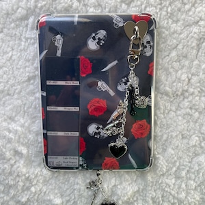 May include: A rectangular phone case with a black background featuring skulls, red roses, and handguns. The case has a clear border and a silver charm with hearts and a sword. Text on the case reads "Black Heart", "Weapon Play", "Dark Desire", and "Light Choking".