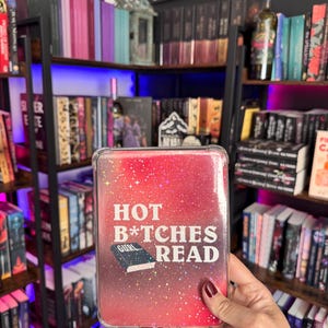 May include: A rectangular e-reader case with a clear, glittery finish over a red and pink gradient background. The front features white text that reads "HOT B*TCHES READ" above a small graphic of a black book with the word "GURL..." on its cover.