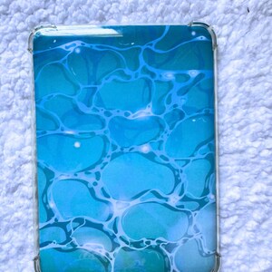 May include: A rectangular protective case for a tablet or electronic device featuring a vibrant blue and turquoise water ripple pattern. The design mimics the appearance of sunlight reflecting on the surface of a pool with light blue lines.