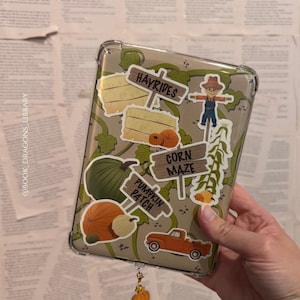 May include: Clear phone case decorated with fall-themed stickers, including pumpkins, hay bales, and a scarecrow. The case has a pumpkin charm attached. The background is a stack of old book pages.
