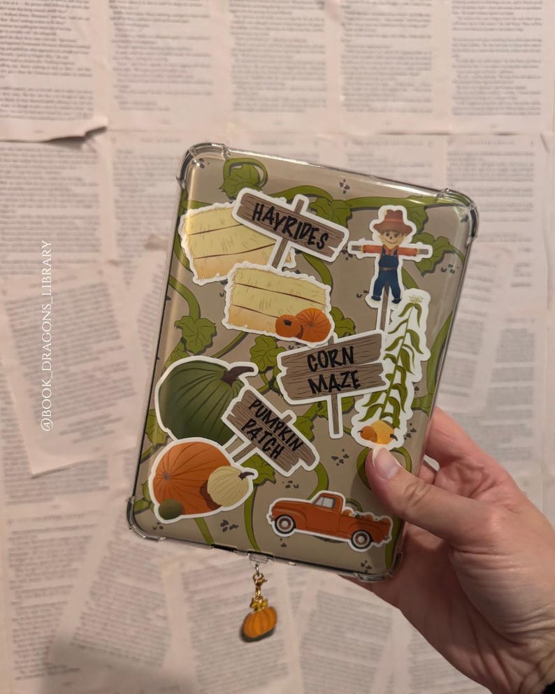 Pu&ograve; includere: Custodia per telefono trasparente decorata con adesivi a tema autunnale, tra cui zucche, balle di fieno e uno spaventapasseri. La custodia ha un ciondolo a forma di zucca attaccato. Lo sfondo &egrave; una pila di vecchie pagine di libri.