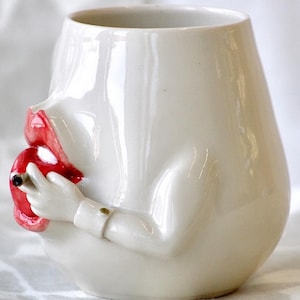 Peut inclure: Une tasse en céramique blanche en forme de visage avec des lèvres rouges et une main tenant une cigarette. La tasse est un objet de fantaisie.
