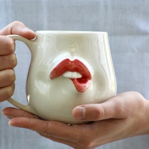 May include: White ceramic mug shaped like a face with a pair of red lips. The lips are slightly parted and the mug has a handle.
