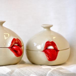Op de afbeelding: Twee keramische containers met een witte glazuur en rode lippen en tong geschilderd op de voorkant. De containers zijn rond en hebben een deksel.