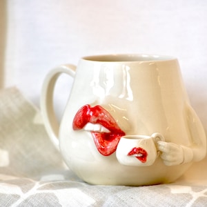 May include: White ceramic mug shaped like a face with red lips and a small white mug in its hand.