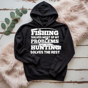 Op de afbeelding: Zwarte hoodie met witte tekst: "FISHING SOLVES MOST OF MY PROBLEMS HUNTING SOLVES THE REST". Het ontwerp bevat een hengel en een geweer. De hoodie heeft een voorzak en een capuchon met trekkoord.