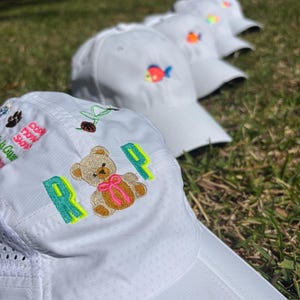 May include: Several white baseball caps with embroidered designs are arranged on green grass. One cap features the letters "R" and "P" in neon green, a teddy bear with a pink bow, and other small embroidered images. Other caps have a fish design.