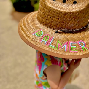 May include: A straw hat with a wide brim, featuring the name "PALMER" embroidered in neon green, pink, and blue. The hat has a dark brown band and three black eyelets on the crown. The hat is worn by a person wearing a colorful dress.