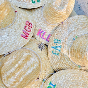 May include: A collection of straw hats with personalized embroidered initials in various colors. Each hat features a natural straw color, a chin strap, and colorful embroidered initials, perfect for a beach day or outdoor event.