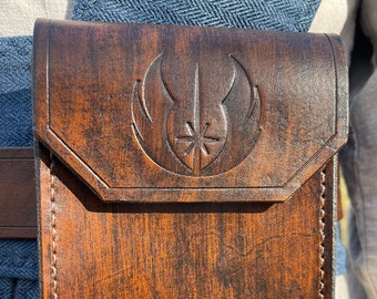 May include: A brown leather pouch with a flap closure, featuring a detailed embossed emblem. The pouch is attached to a brown belt and has visible stitching along the edges. The leather has a distressed finish, giving it a vintage look.