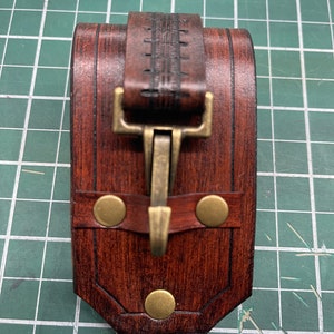 May include: A handcrafted, rectangular leather case in shades of brown, featuring a decorative strap and a brass clasp. The case is secured with three brass rivets and is designed with a detailed, textured finish.