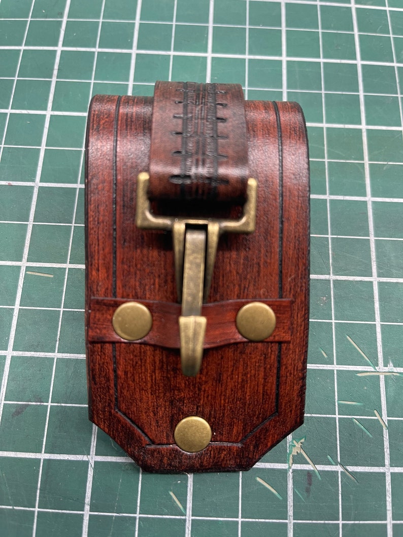 May include: A handcrafted, rectangular leather case in shades of brown, featuring a decorative strap and a brass clasp. The case is secured with three brass rivets and is designed with a detailed, textured finish.