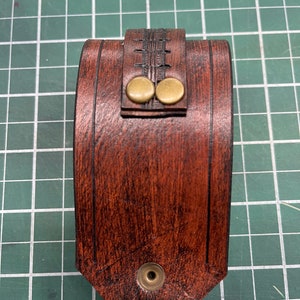 May include: A handcrafted, brown leather wrist cuff with a decorative stitched design. The cuff features two brass-colored button closures and a single snap closure. The leather has a wood-grain appearance.