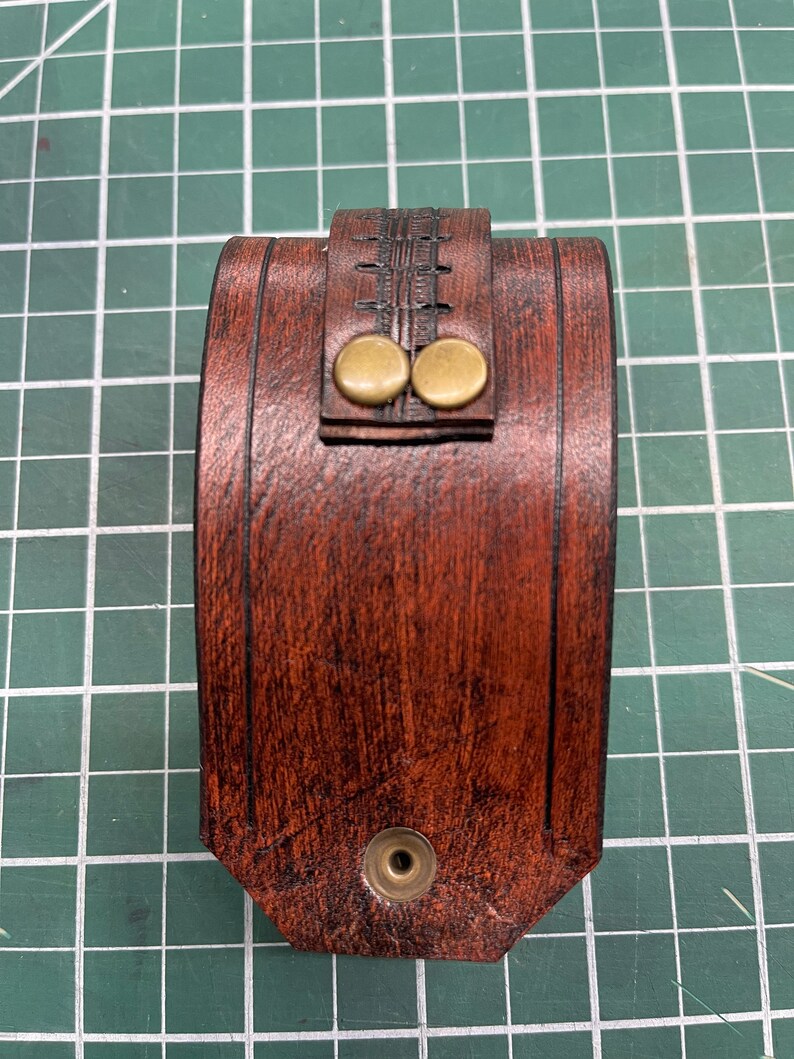 May include: A handcrafted, brown leather wrist cuff with a decorative stitched design. The cuff features two brass-colored button closures and a single snap closure. The leather has a wood-grain appearance.