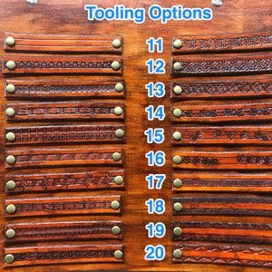 May include: A wooden display board with the text "Tooling Options" features twenty samples of tooled leather straps. Each strap is a different design, ranging from simple lines to intricate floral patterns. The straps are secured with brass rivets.