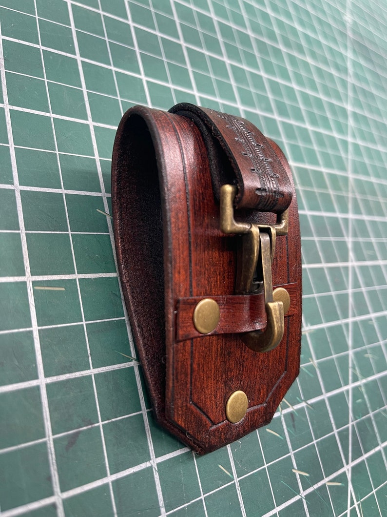 May include: A handcrafted, dark brown leather pouch with a brass clasp and rivets. The pouch has a rectangular shape with a curved top and is designed to be worn on a belt. The leather has a rich, textured appearance.
