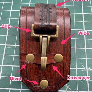 May include: A handcrafted, dark brown leather bracelet with a brass-colored clip and rivets. The wide band features a decorative accent and a crossover design, showcasing a rustic aesthetic.