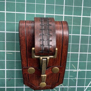 May include: A handcrafted, dark brown leather pouch with a decorative stitched strap and brass-colored clasp. The pouch features a rectangular shape with rounded edges and two brass rivets. The leather has a rich, textured appearance.