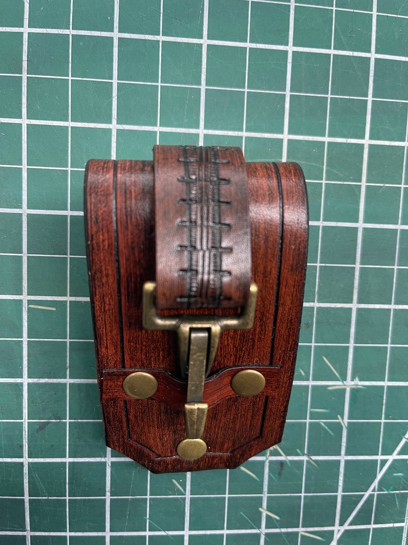 May include: A handcrafted, dark brown leather pouch with a decorative stitched strap and brass-colored clasp. The pouch features a rectangular shape with rounded edges and two brass rivets. The leather has a rich, textured appearance.