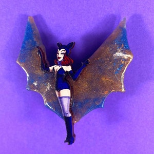 Peut inclure: Une broche en résine de chauve-souris bleue et dorée à paillettes avec une femme de dessin animé en costume bleu et noir.