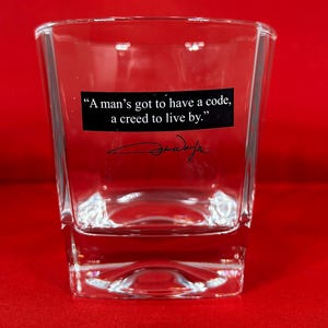 May include: Clear square shot glass with the quote "A man's got to have a code, a creed to live by." in black text on a black rectangle. The glass also features a signature. The background is red.