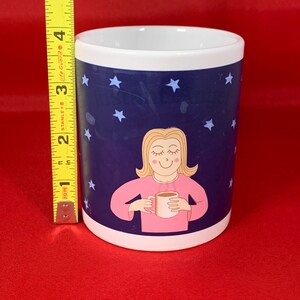 K&ouml;nnte beinhalten: Eine wei&szlig;e Keramiktasse mit blauem Hintergrund und wei&szlig;en Sternen. Eine Comic-Frau mit blonden Haaren h&auml;lt eine Tasse Kaffee und l&auml;chelt. Der Text auf der Tasse lautet "Ich sage nicht, dass ich faul bin, aber ich sage auch nicht, dass ich nicht faul bin."