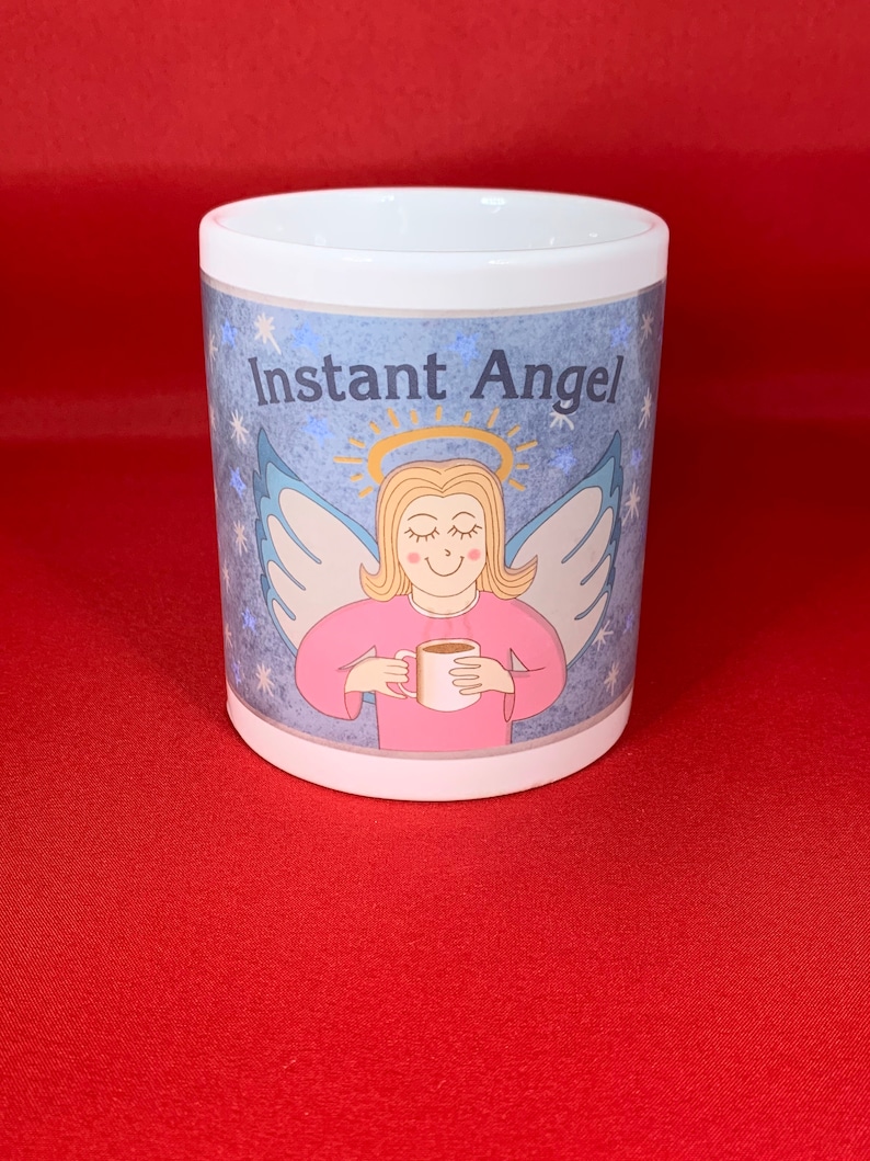 K&ouml;nnte beinhalten: Wei&szlig;e Keramiktasse mit einer blauen und rosa Illustration eines Engels, der eine Tasse Kaffee h&auml;lt. Der Text "Instant Angel" ist auf die Tasse gedruckt.