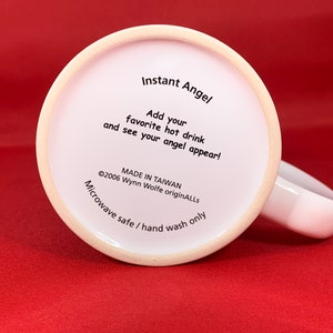 K&ouml;nnte beinhalten: Wei&szlig;e Keramiktasse mit schwarzem Text, der "Instant Angel" und "F&uuml;gen Sie Ihr Lieblingsgetr&auml;nk hinzu und sehen Sie, wie Ihr Engel erscheint!" lautet. Der Text enth&auml;lt auch "&copy;2006 Wynn Wolfe originALLS" und "MADE IN TAIWAN". Der Boden der Tasse lautet "Mikrowellengeeignet / Nur Handw&auml;sche".