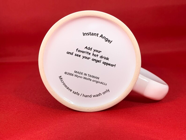 K&ouml;nnte beinhalten: Wei&szlig;e Keramiktasse mit schwarzem Text, der "Instant Angel" und "F&uuml;gen Sie Ihr Lieblingsgetr&auml;nk hinzu und sehen Sie, wie Ihr Engel erscheint!" lautet. Der Text enth&auml;lt auch "&copy;2006 Wynn Wolfe originALLS" und "MADE IN TAIWAN". Der Boden der Tasse lautet "Mikrowellengeeignet / Nur Handw&auml;sche".