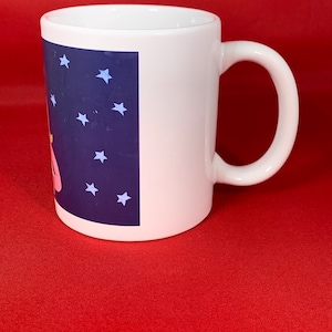 K&ouml;nnte beinhalten: Wei&szlig;e Keramiktasse mit einem blauen und wei&szlig;en Design mit Sternen und einer rosa Figur. Die Tasse hat einen Henkel.