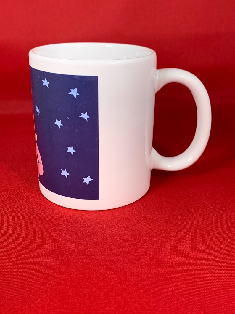 K&ouml;nnte beinhalten: Wei&szlig;e Keramiktasse mit einem blauen und wei&szlig;en Design mit Sternen und einer rosa Figur. Die Tasse hat einen Henkel.
