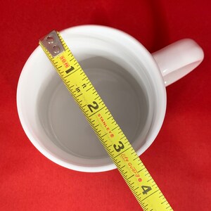 K&ouml;nnte beinhalten: Wei&szlig;e Keramiktasse mit einem gelben Ma&szlig;band darum gewickelt. Das Band misst 10 cm.