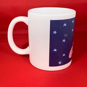 K&ouml;nnte beinhalten: Wei&szlig;e Keramiktasse mit einem blauen und wei&szlig;en Design mit einer Comicfigur und Sternen. Die Tasse hat einen Henkel.