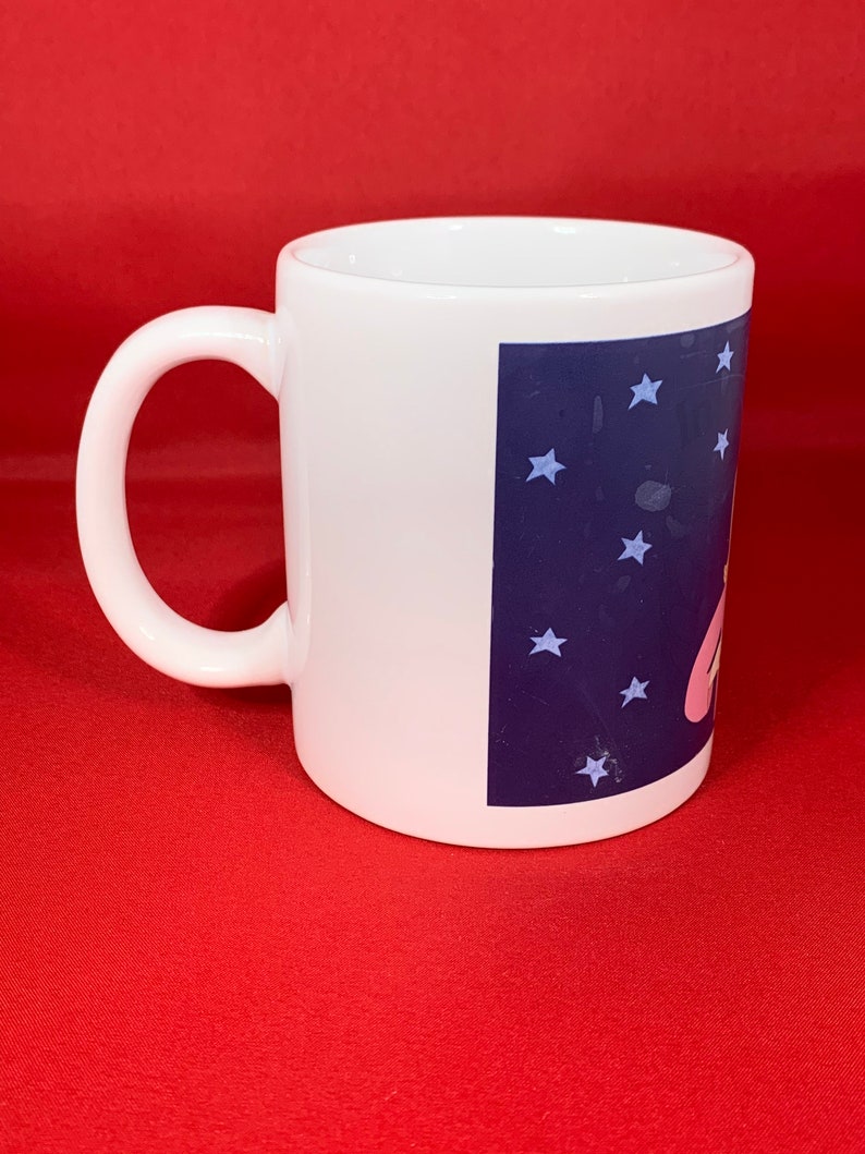K&ouml;nnte beinhalten: Wei&szlig;e Keramiktasse mit einem blauen und wei&szlig;en Design mit einer Comicfigur und Sternen. Die Tasse hat einen Henkel.