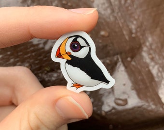 May include: A small, die-cut sticker featuring a cartoon puffin. The bird has a black back, white belly, and a large, colorful beak with orange, yellow, and red hues. The puffin has a purple eye and orange feet.