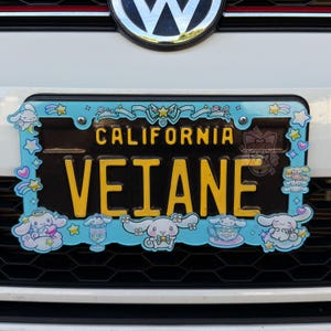 Puede incluir: Una placa de matr&iacute;cula de California con el nombre "VEIANE" en letras amarillas. La placa est&aacute; rodeada por un marco azul claro con personajes de dibujos animados y la palabra "CALIFORNIA" en amarillo.