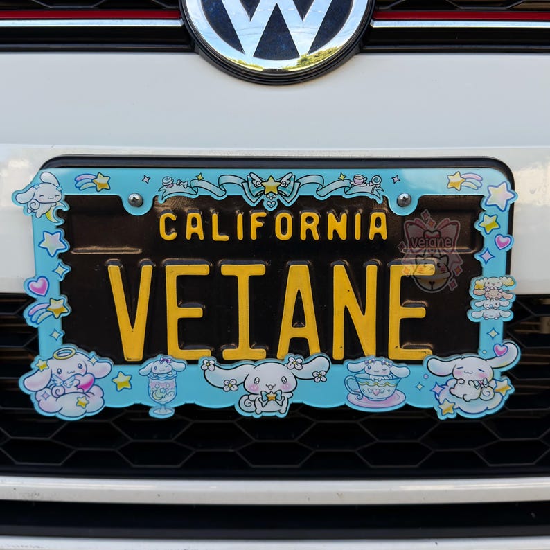Puede incluir: Una placa de matr&iacute;cula de California con el nombre "VEIANE" en letras amarillas. La placa est&aacute; rodeada por un marco azul claro con personajes de dibujos animados y la palabra "CALIFORNIA" en amarillo.